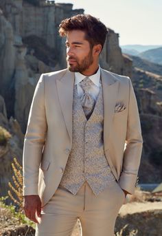 a man wearing a suit and tie standing in front of a mountain range with his hands in his pockets