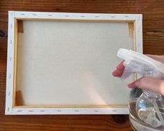 a person holding a spray bottle in front of a white frame on a wooden table