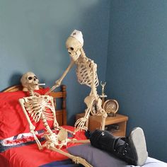 two skeletons sitting on a bed in a room with blue walls and red sheets, one is laying down