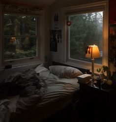 an unmade bed in a dimly lit room with two windows and a lamp on the nightstand