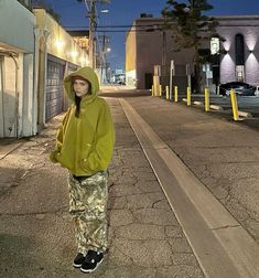 a person standing in the middle of an empty street at night wearing a yellow hoodie and camo pants