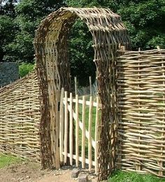 a wooden fence made out of branches and sticks with an arch in the middle, surrounded by grass