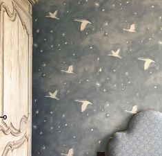 a room with a bed, chair and wallpaper that has white birds flying in the sky