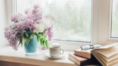 an open book and cup on a window sill next to a vase with flowers