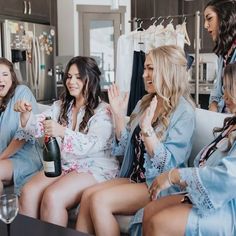 four women in robes sitting on a couch and one is holding a bottle of champagne