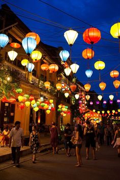 many people are walking down the street at night with colorful lanterns hanging from the ceiling