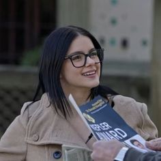 a woman in glasses is holding a book and talking to another person wearing a coat