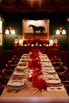 a long table set with place settings and red flowers on the runneres in front of a horse painting