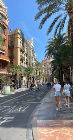 two people walking down the street in front of some buildings and palm trees on a sunny day