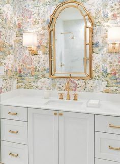 a bathroom with floral wallpaper and gold accents on the mirror above the double sink