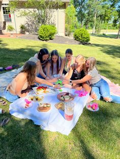 a group of women sitting around a table with food on it in the grass outside