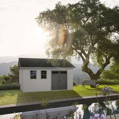 a small white shed sitting next to a tree on top of a lush green field