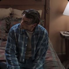 a man sitting on top of a bed next to a night stand with a lamp