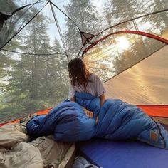 a woman sitting on top of a sleeping bag in a tent with trees in the background