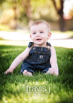 a baby sitting in the grass with his eyes open and smiling at the camera while wearing overalls