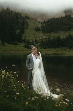 a bride and groom standing next to each other near a lake in the mountains on their wedding day