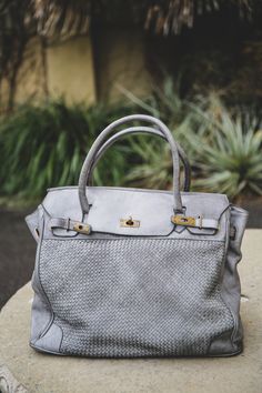 The Rosalind tote in grigio (gray). Front view sitting on tabletop. Vintage, soft woven leather with brass clasp & lock. Luxury Satchel Tote For On-the-go, Luxury Weekender Bag For On-the-go, Luxury Satchel With Rolled Handles For On-the-go, Luxury Tote Satchel For On-the-go, Chic Satchel Travel Bag For On-the-go, Top Handle Weekender Bag For Shopping, Trendy Travel Bag With Rolled Handles, Chic On-the-go Satchel Travel Bag, Chic Satchel With Braided Handles In Tote Shape