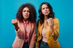 two women standing next to each other in front of a blue background with one pointing at the camera