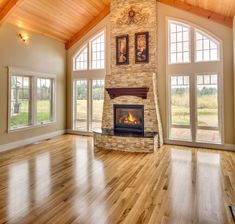 an empty living room with wood flooring and stone fire place in the center surrounded by large windows