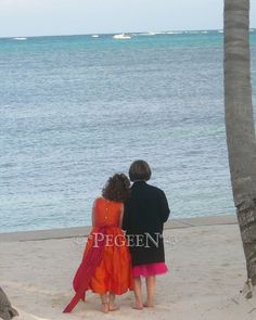 two people are standing on the beach looking out at the water and palm trees in front of them