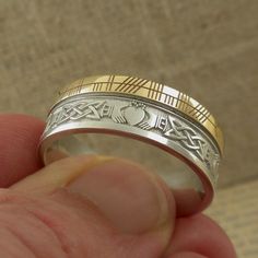 Sterling Silver and 10K Yellow Gold with Claddagh and Celtic Knot Design around the entire ring. A custom personalized phrase in Ogham on 10K yellow rail edge. Claddagh & Celtic Knot Wedding Band with Ogham Script Rail. Handmade in Ireland. 7.5 mm wide. Available in whole & half Sizes 4.5 to 13. Florentine background with polished design, your personalized phrase in Ogham Script on rail edge. Comfort Fit. Irish Hallmark. The phrase is usually a name or word or short phrase. Start a conve Ceremonial Silver Engraved Ring Stamped 14k, Ceremonial Silver Engraved Ring, Stamped 14k, Unique Engraved Yellow Gold Wedding Ring, Symbolic Hallmarked Wedding Rings, Symbolic Wedding Rings Hallmarked, Symbolic Polished Wedding Jewelry, Symbolic Wedding Jewelry With Polished Finish, Hallmarked Jewelry For Wedding, Symbolic Polished Jewelry For Wedding