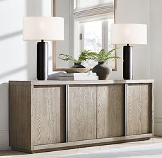 two lamps sitting on top of a wooden cabinet in front of a window with a potted plant