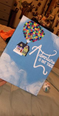 a person holding up a painting that says adventure is out there with a house and hot air balloon in the sky