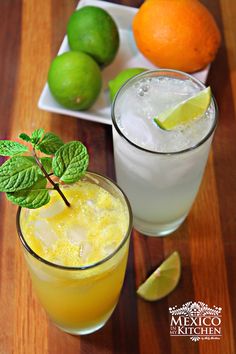 two glasses filled with drinks sitting on top of a wooden table next to limes and oranges