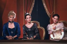 three women sitting at a table in front of a red curtain, one wearing a tiara