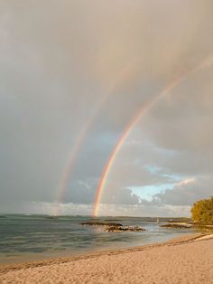 two rainbows in the sky over a beach with water and trees on it's side