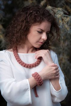 a woman wearing a red necklace and white shirt standing in front of some trees with her hands on her chest