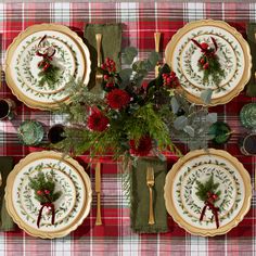 the table is set for christmas dinner with red and green plates, napkins, silverware and greenery