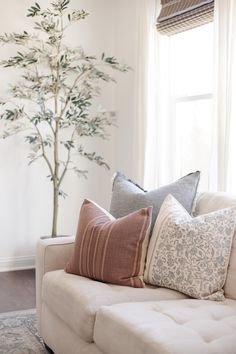 pillows on a couch in front of a window with a potted tree behind it