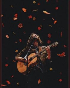 a man holding a guitar and singing into a microphone with autumn leaves falling around him