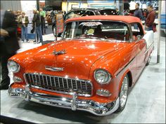 an orange classic car on display in a showroom with people looking at the cars