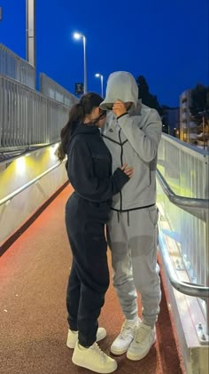 two people standing next to each other on a bridge at night with lights in the background