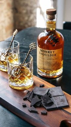 a wooden tray topped with two glasses filled with liquid next to a bottle of whiskey