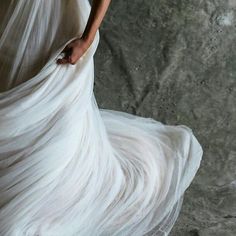 a woman in a long white dress is standing near a rock wall with her hands on the skirt