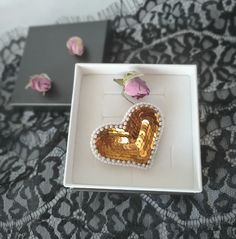 a heart shaped brooch sitting on top of a white box next to pink flowers