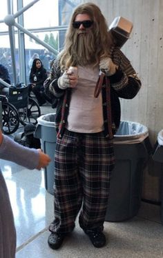 a man with long hair and beard standing in front of a trash can wearing plaid pants