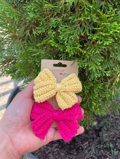 a hand holding two crocheted bows in front of a plant with a tag on it