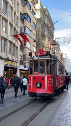 a red trolley car traveling down a street next to tall buildings and people walking on the sidewalk