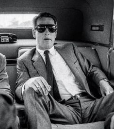 two men sitting in the back of a car wearing suits and ties, one with sunglasses on