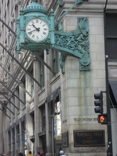 a large clock mounted to the side of a building on a city street with people walking by