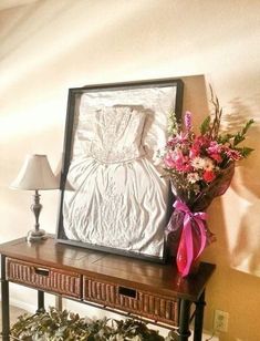 a vase with flowers sitting on top of a wooden table next to a framed dress