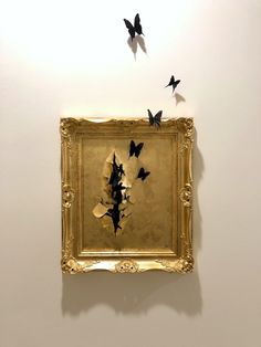 several black butterflies are flying in the air above a gold framed artwork on a white wall