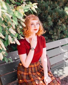 a woman sitting on top of a wooden bench next to trees and bushes with her hands under her chin