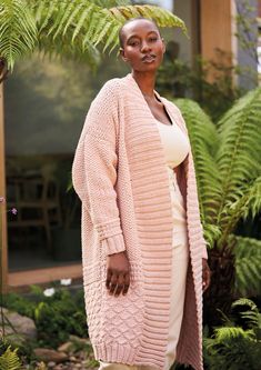 a woman standing in front of some plants wearing a pink cardigan and tan pants