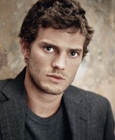 a man with curly hair wearing a suit and looking at the camera while standing in front of a wall