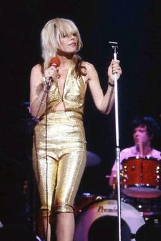 blond haired woman in gold outfit holding a microphone and singing into a mic on stage